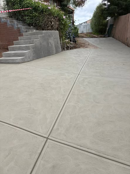 Broom-finished concrete driveway with feature joint lines running past a brick retaining wall and matching concrete steps